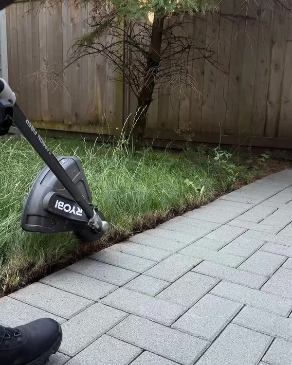 Close-up of a powered trimmer clearing an overgrown edge beside an interlocking paver walkway